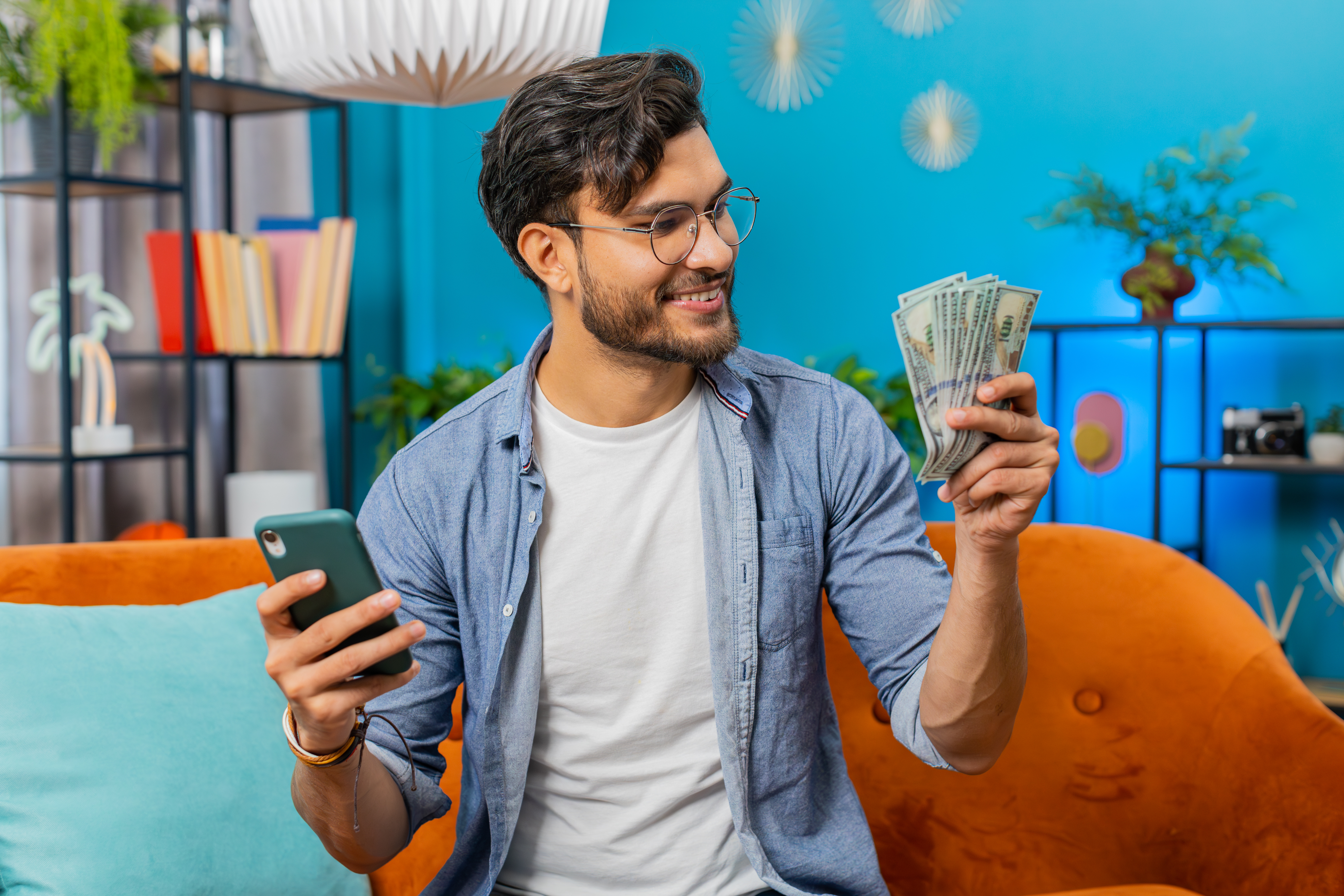 Planning budget. Rich happy Indian man use smartphone calculator app count money cash, calculate domestic bills at home. Guy satisfied of income earnings win saves money for planned vacation gifts