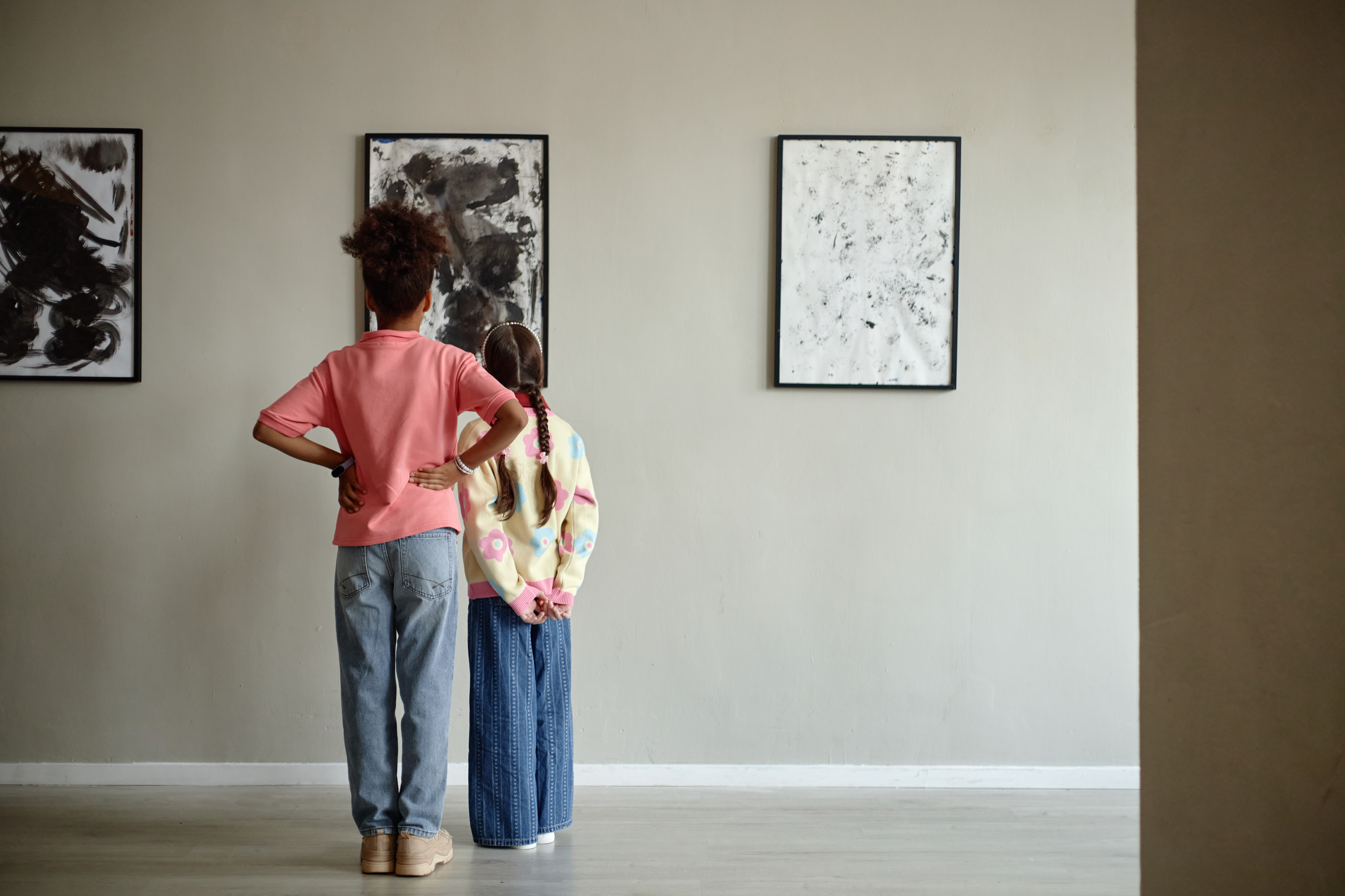 Diverse children standing back to camera, looking at paintings in art gallery. Image represents early childhood education and cultural development, ideal for school curriculum promotion