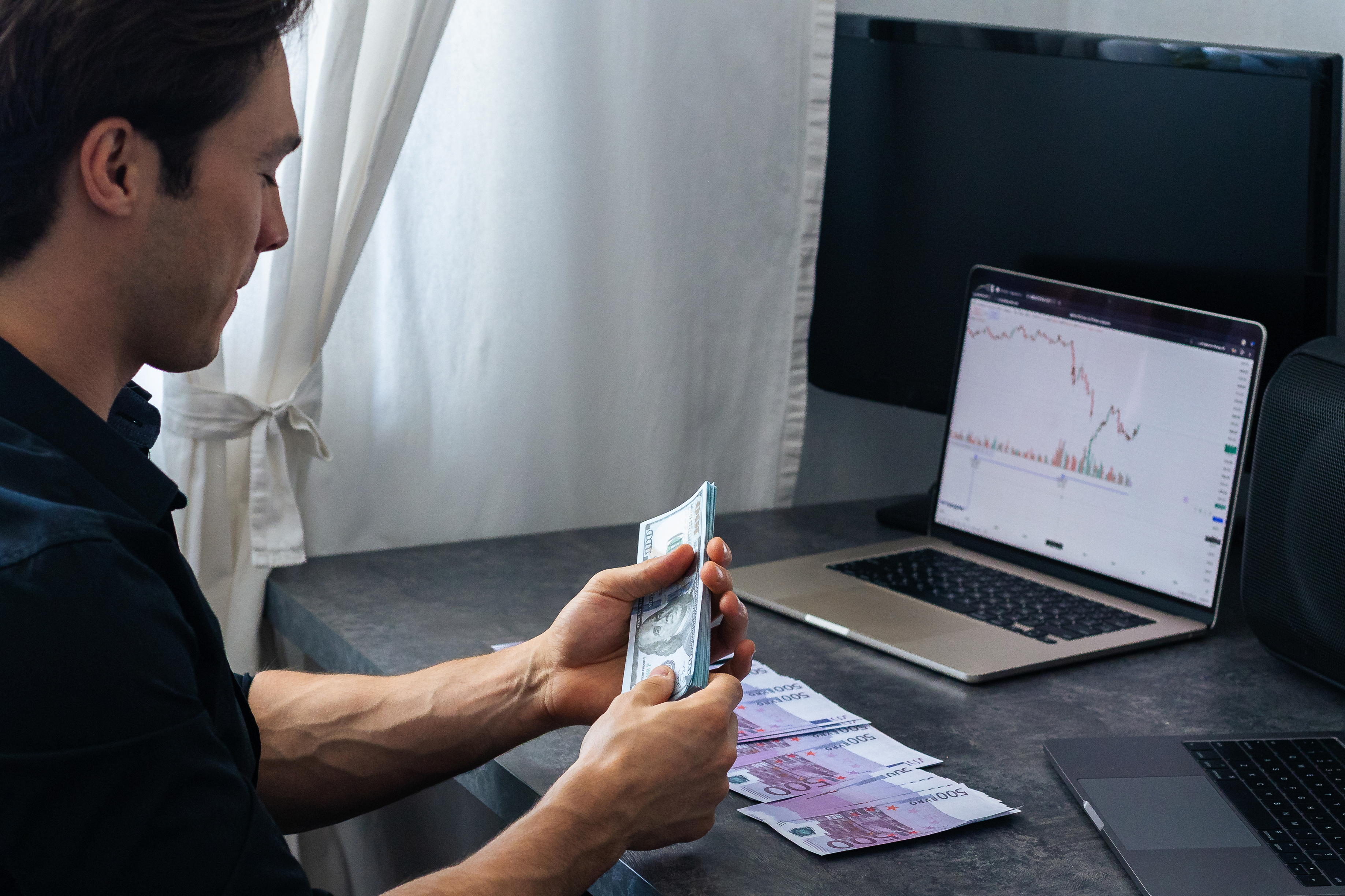 Man examines cash in hand while reviewing financial charts on a laptop, with banknotes spread out on the desk in a contemporary workspace